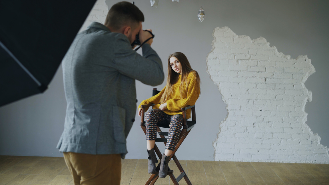 Young beautiful woman model posing for photographer while he is shooting with a digital camera in photo studio indoors